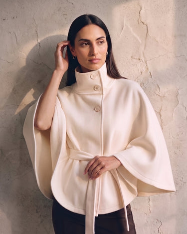 The Lorenne cape coat in white — on model with hand raised to her hair, showing the cape drape