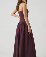 A woman with long brown hair models the Tamsin Strapless Dress, a floor-length plum evening party dress. She stands sideways, looking over her shoulder against a plain light gray background.