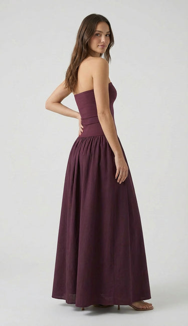 A woman with long brown hair models the Tamsin Strapless Dress, a floor-length plum evening party dress. She stands sideways, looking over her shoulder against a plain light gray background.