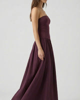 A woman stands in profile against a light gray background, wearing the Tamsin Strapless Dress—a high-waisted, floor-length dark purple evening gown—paired with nude sandals.