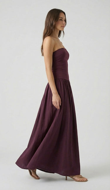 A woman stands in profile against a light gray background, wearing the Tamsin Strapless Dress—a high-waisted, floor-length dark purple evening gown—paired with nude sandals.
