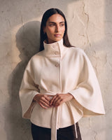 The Lorenne cape coat in white — front view on model, hands clasped at the belted waist