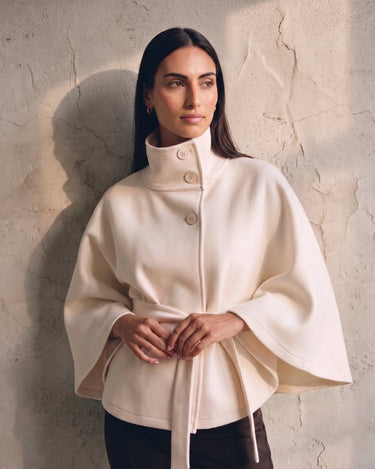 The Lorenne cape coat in white — front view on model, hands clasped at the belted waist