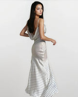 A woman with long dark hair models the Elowen Polka Dot Dress, a sleeveless, floor-length chiffon A-line dress, standing against a plain light background and glancing over her shoulder.