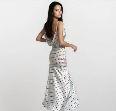 A woman with long dark hair models the Elowen Polka Dot Dress, a sleeveless, floor-length chiffon A-line dress, standing against a plain light background and glancing over her shoulder.