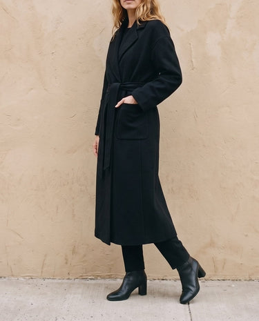 The Caroline Tie Waist Coat in black — longline belted coat, front view with hand in pocket on model