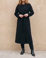 The Caroline Tie Waist Coat in black — longline belted coat, front view tying the waist belt on model