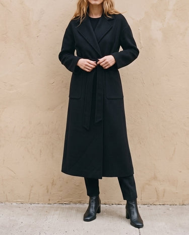 The Caroline Tie Waist Coat in black — longline belted coat, front view tying the waist belt on model