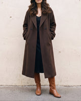 The Chocolate Maxi Coat in chocolate brown — longline coat, front view with hands in pockets on model