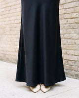 Detail of the lower half of the black Eclipse Satin Dress showing the flared floor-length hem