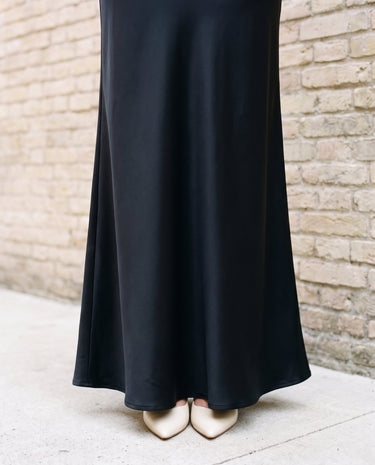 Detail of the lower half of the black Eclipse Satin Dress showing the flared floor-length hem