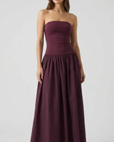A woman stands against a plain background wearing the Tamsin Strapless Dress in dark purple, featuring a fitted bodice and gathered, floor-length skirt. Her long hair is loose, and she is barefoot.
