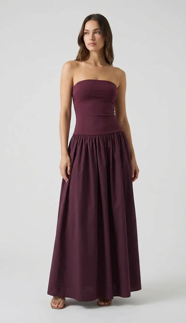 A woman stands against a plain background wearing the Tamsin Strapless Dress in dark purple, featuring a fitted bodice and gathered, floor-length skirt. Her long hair is loose, and she is barefoot.