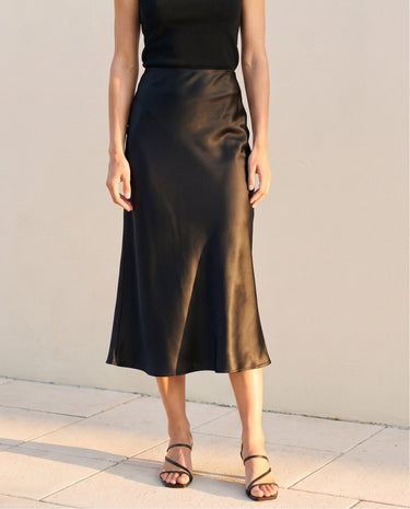 Model wearing black satin midi skirt with a black sleeveless top and strappy sandals, front view – Moriel Heritage