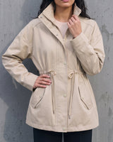 Model wearing the light khaki Halden Coat with hands on hips, front view showing the drawstring waist