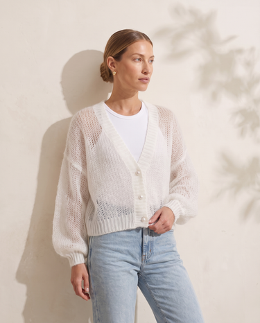 Marisol Knit Cardigan in White, model leaning against a limewash wall showing the balloon sleeves