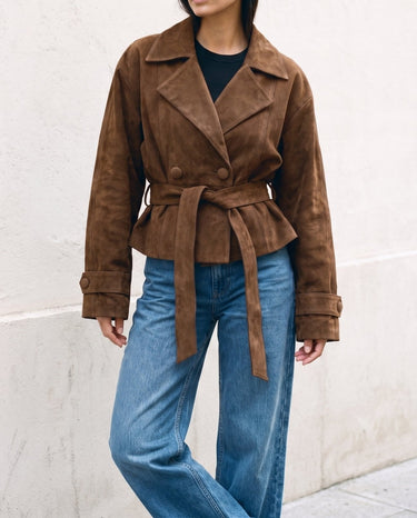 Model wearing the dark brown Margot Tie-waist Coat tied at the waist, front view against a stone wall