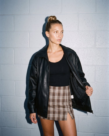 Model holding open the black Marseille Faux Leather Jacket, front view over a black top and check mini skirt