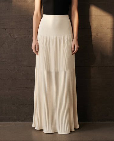 Model wearing the ivory Matilde Skirt, front view showing the smooth yoke over the hip and pleats falling to a floor-length hem, styled with a black top