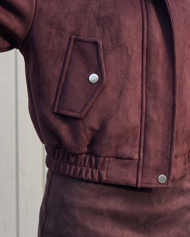 Close-up of the bomber jacket's press-stud flap pocket and elasticated hem in burgundy suede-look fabric