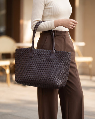 The Amalfi Woven Tote in brown carried by hand beside chocolate trousers at a cafe terrace