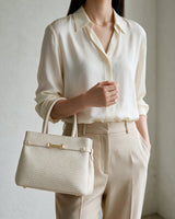 The Camille Handbag in ivory carried by hand with a cream shirt and beige trousers