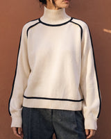 Model wearing cream high-neck knit sweater with grey trousers, hands in pockets – Moriel Heritage