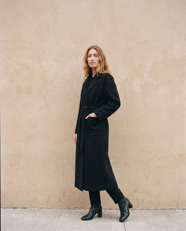 The Caroline Tie Waist Coat in black — longline belted coat, front view with hand in pocket on model