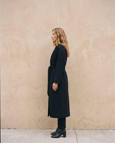 The Caroline Tie Waist Coat in black — longline belted coat, side view on model