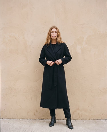 The Caroline Tie Waist Coat in black — longline belted coat, front view tying the waist belt on model