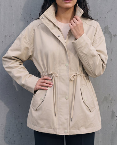 Model wearing the light khaki Halden Coat with hands on hips, front view showing the drawstring waist