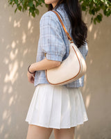 The Ines Crescent Bag in cream carried on the shoulder with a blue checked shirt and white pleated skirt
