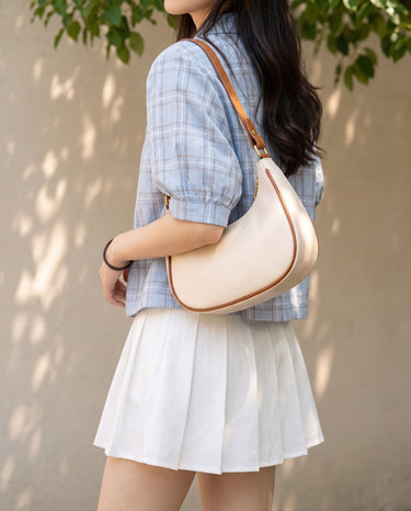 The Ines Crescent Bag in cream carried on the shoulder with a blue checked shirt and white pleated skirt