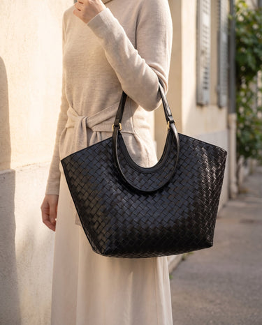 The Lucca Woven Tote in black carried on the shoulder with a cream knit and long skirt on a city street