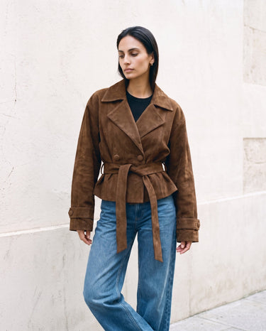 Model wearing the dark brown Margot Tie-waist Coat tied at the waist, front view against a stone wall