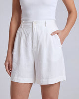 The Mauria Linen Shorts in white — pleated linen shorts with side pockets, hand-in-pocket view on model