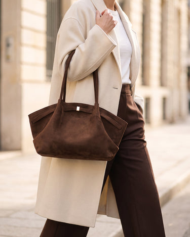 The Odette Suede Tote in brown carried over the shoulder with a cream wool coat and chocolate trousers on a sunlit street