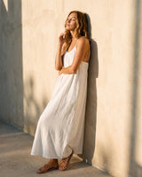 The Rylie Dress in white — flowing maxi dress, front view on model leaning against wall