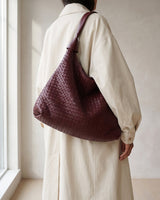 The Siren Woven Bag in burgundy carried on the shoulder with a cream coat